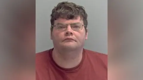 Police custody photograph of Daniel Russell. He is wearing glasses and is  looking at the camera. He is wearing a red t-shirt.