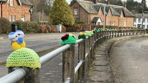 Andrew Findlay A curved roadside railing is decorated with colorful knitted hats and small crochet figures, forming a playful line along a quiet street. Brick houses and trees sit in the background, giving the scene a cosy village feel.