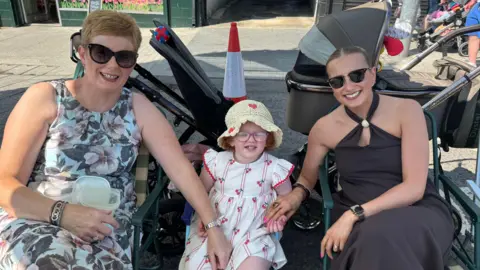Ellen Watson has light brown hair tied back an parted in the middle and is wearing sunglasses. A middle aged woman with short reddish hair and sunglasses is on the left and a young girl with glasses and a sun hat is in the middle