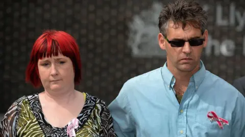 Getty Images Photograph of Paul Jones (right) in 2013. He wears a blue shirt with a pink breast cancer bow on his shirt pocket.His wife and April's mum, Coral (left), has bright red and black hair in a bob. She wears a black and white zebra blouse. 