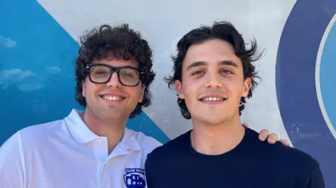 Vincenzo, on the left with dark curly hair and glasses, wearing a white supporters' club polo shirt, smiles beside his friend Francesco, in a navy t-shirt. He also has shaggy dark hair and is smiling for the photo.