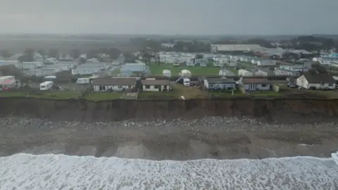 Waves break on a beach in front of brown muddy cliffs. There are several bungalows and dozens of caravans on the cliff top, some are close to the edge.