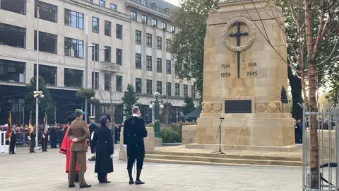 A group of people in official dress approach the Cenotaph. In the distance crowds can be seen looking on behind metal barriers. 