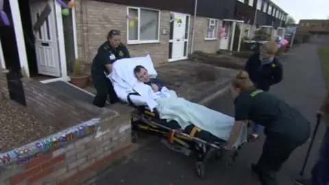 Ambulance staff wheel Mr Dobbin, who is on a stretcher, into his terrace house. His house and neighbouring homes have balloons and banners on them.