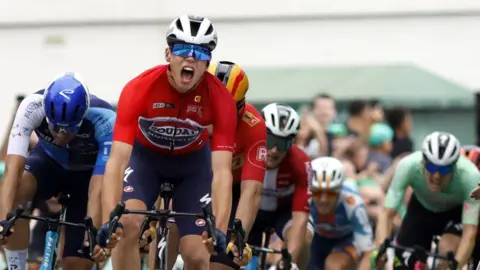 Cyclists all wearing helmets and colourful jerseys race in front of a crowd. The man leading the race has his mouth open as if he is shouting,