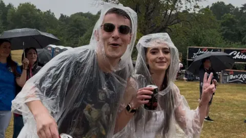 Man and woman wearing clear raincovers. Man wears sunglasses. Woman shows the peace sign with her fingers. They are smiling and walking across a field. Two women are in the background with an umbrella covering their heads from the rain. All are in a field where a music festival is being held. 