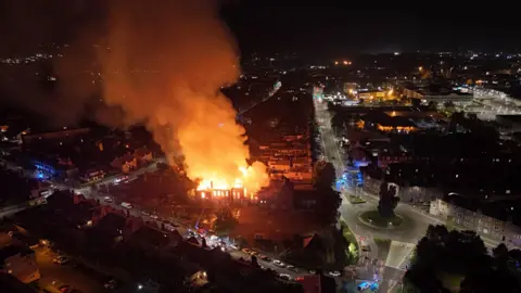 STUART COWPER An aerial shot of the fire at the former primary school. A large column of orange smoke can be seen blowing over the city. The school is glowing orange with fire below.