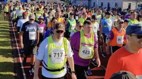 The first colored photo of the article. Shows lots of people on an athletics track. Everyones tops are different colours, and some wear high viz bibs. 