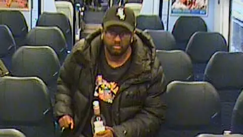 British Transport Police Zakaria Mohammed, wearing a black Boston Red Sox cap, black coat and black T-shirt with a red and yellow pattern on it, walking through the empty train.