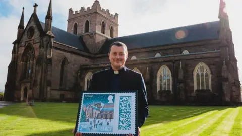 Royal Mail The Dean of Armagh, The Very Reverend Shane Forster, holding a small sign with the image of the stamp design on it - the stamp is an illustration of St Patrick's Cathedral with lots of children outside of it playing in the snow. The cathedral is covered in snow with the lights on and the sky has a dark sunset. The Dean is standing outside the cathedral while holding the stamp. 