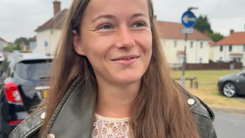 BBC News A young mum in a black leather jacket and long brown hair smiles nervously while looking off camera.