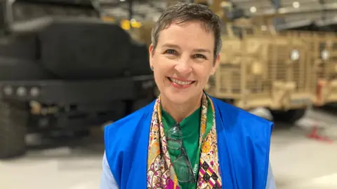 A woman wearing a green top and multicoloured scarf smiles into the camera. Behind her are the blurred outlines of a grey and khaki vehicle.