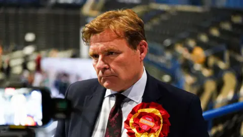 A man with ginger hair looking solemn, wearing a red and yellow 'Vote Labour' rosette. 