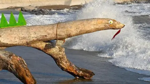 Mike Pugh Lizzy the lizard, actually a piece of driftwood, washed up on a beach in Porthcawl. The driftwood has white goggly eyes attached to the front of it, a red tongue, and has a wooden plaque necklace which reads "Lizzy". Green spikes can also be seen on its back. 
