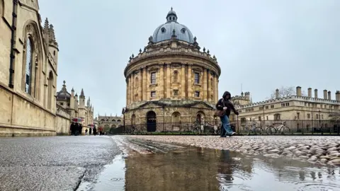 BBC Weather Watcher EstherJ Oxford - paths and cobbled streets with puddles dotted about buildings in the background and people walking with hoods and umbrellas up.