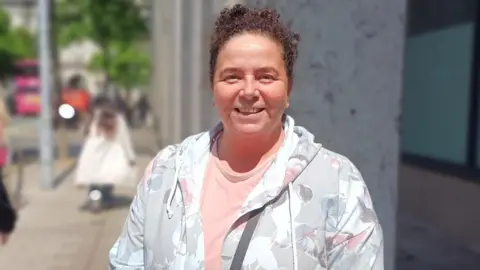 A woman is standing in a street looking into the camera. Her curly hair is pulled back. She's wearing a pink top and a floral coat.
