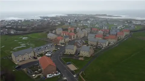 An aerial shot of a modern housing estate called The Kilns which is cluster of 45 modern houses. Beyond them is the rest of the village of Beadnell and in the far distance the beach and the sea. The Kilns is bordered on the left and right with grassy open space.
