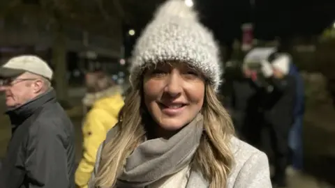 A woman with long blonde hair, a white bobble hat and a white coat in the dark