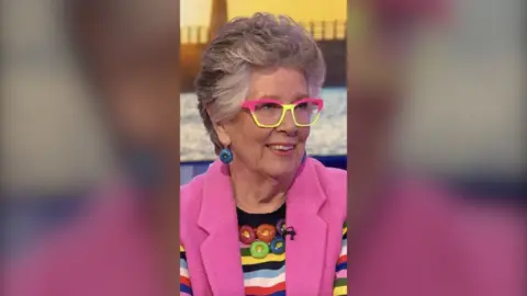 Prue Leith wears pink and yellow glasses, and a multicoloured striped top under a pink sleeveless cardigan