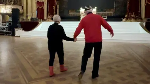 An elderly man and woman hold hands as they dance in a ballroom. 