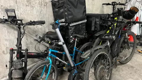 O bicicletă albastră se află în față, printre alte două biciclete negre și o trotinetă, unele având cutii mari prinse pe ele, precum și mănuși pe mânere.