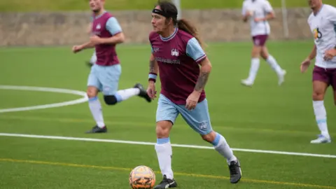 A wide shot of Steve Harris dress in a Claret shirt and light blue shorts, which have Iron Maiden printed across the chest. He has long brown hair tied back and had an orange football at his feet.