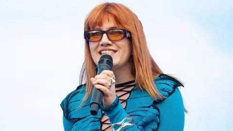 CMAT, she is looking to her right while holding a microphone, she is wearing a blue tp[ and has red hair. She is wearing sunglasses.