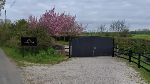 Google The entrance to The Old Essex Barn wedding venue. There are a pair of large black wooden gates across a grey gravel driveway, with a picket fence to the right hand side, and behind the gate can be seen green fields and hedgerows and a pink blossom tree. There is a black sign to the left of the gate which says Old Essex Barn.