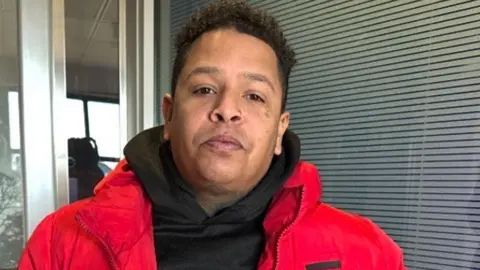 A man with black curly hair, a black top and bright red coat in front of a window with blinds drawn