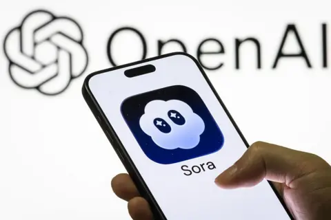 Getty Images A person holding a phone with the Sora logo. It is a cloud with eyes visible on the screen against a white backdrop with the OpenAI logo displayed.