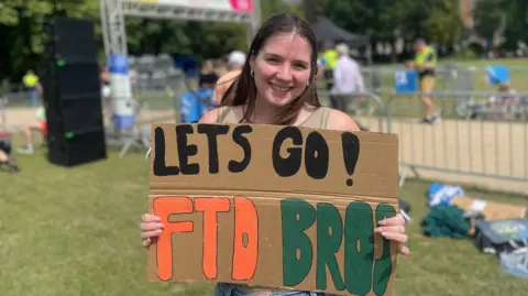 A smiling woman stands outdoors at what looks like a public event, holding a handmade cardboard sign that reads: “LETS GO! FTD BROS” in large black, orange, and green letters. She has dark hair and is standing in front of metal barriers, with people and trees out of focus in the background on a sunny day.