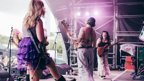 Derek Bremner Panic Shack performing connected  signifier    astatine  a festival. Megan, pictured connected  the right, smiles wide    astatine  her bandmates portion    holding her guitar. Bassist Emily Smith and guitarist Romi Lawrence are pictured with their backs to the camera portion    vocalist  Sarah Harvey is astatine  the apical  of the signifier    singing to the crowd. 