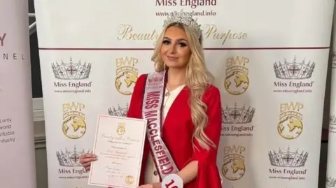 A woman wearing a white dress with a red jacket is smiling holding a certificate, wearing a Miss Macclesfield sash. She is standing in-front of a sign saying Miss England that is covered in crowns. 