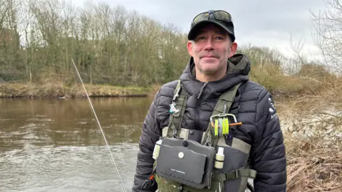 Del Spry standing in front of a river. He is wearing a black jacket, cap and sunglasses on his head and a khaki-coloured chest pack with fishing gear attached to it. The riverbank on the opposite side is lined with bare trees.