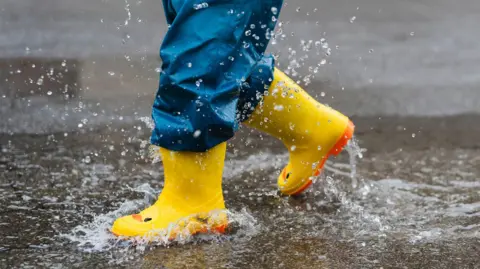 A child walking through a puddle in yellow wellington boots which have a face on them to make them look like a duck.
