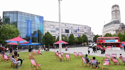 Alex Pope People sit on deckchairs on the grass outside the entrance to the Luton Point shopping centre. A red Routemaster bus is parked next to a police van on a paved area. The clock tower of the white Luton Town Hall building is behind the bus.