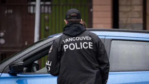 A Vancouver police officer outside a stopped vehicle in Vancouver, British Columbia, Canada, on Wednesday, Sept. 25, 2024. The vehicle is blue. The officer's back is to the camera, and he is wearing a black hooded rain jacket with the words 'VANCOUVER POLICE' on the back written in white.