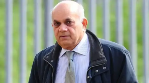 Dr Krishna Singh outside court. He has a bald head and is wearing a blue and white striped shirt and and a grey tie with a blue jacket over the top. He is looking at the camera and there are railings behind him.