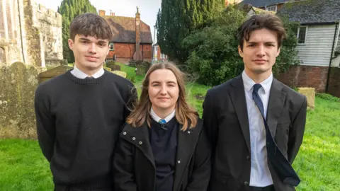 Ian Palmer/ BBC Three teenagers stand looking into the camera. The boy on the left hand side has brown hair and is wearing a white shirt and black jumper. The girl in the middle has long brown hair. She is wearing a black coat, a white shirt, a black jumper and a blue tie. The boy on the right hand side is wearing a black blazer, navy blue tie and a white shirt. He has brown hair.