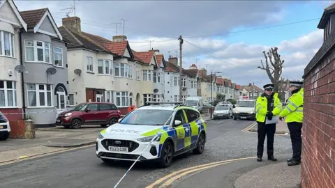 A police car by a taped off area in Southend, showing a brick wall to the right, a road, with cars parked on it, police in the distance and a row of houses to the left. There are also houses to the right. Several trees and a telegraph pole are in the road, and double yellow lines. Two police officers are to the right.