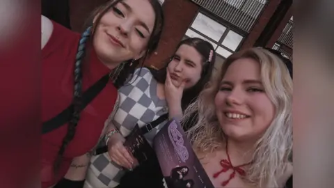 Kirsten Gibson A photo of three young women smiling at the camera for a selfie. The woman on the left is wearing a sleeveless red top with brown and dyed hair in two plaits; in the middle, the woman is wearing a grey and white checked jumper and is cupping her cheeks. She has long brown hair and has a pink clip in. On the right, the woman has shoulder length blonde hair and is wearing a black hat. She can be seen wearing a red bow necklace and is holding a small purple banner.
