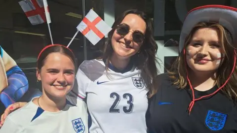 Three young women in England shirts smile at the camera
