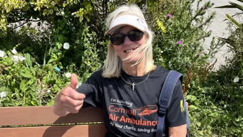 A woman wearing sunglasses, a cap and a 'Cornwall Air Ambulance' t-shirt holds a thumbs up to the camera while sitting on a bench in the sun.
