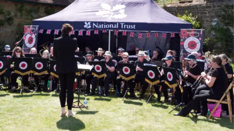 National Trust Images/ Howard Goodengough The image shows an orchestra playing outdoors, with National Trust written on a marquee some players are underneath.