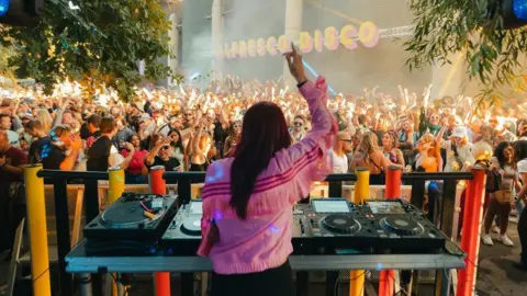 Here and Now A DJ waves her arms in front of a large crowd dancing at an Alfresco Disco event in Bristol