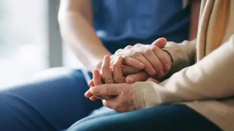 Two people holding hands. One of them wears a cream jumper while the other is wearing blue scrubs. 