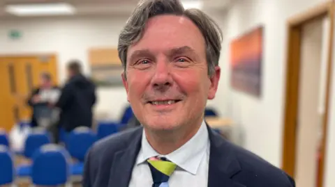 Deputy Andy Sloan - A man with black and grey hair, smiling at the camera with a blue suit on, wearing a white shirt and a multi coloured tie. 
