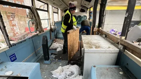 Gloucester City Council Council workers wearing yellow hi-vis vests clearing debris from the inside of the abandoned bus. The vehicle is blue on the inside and filthy, with torn duvets, rubbish and smashed window glass everywhere. 