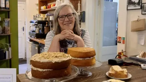Lizzie Leigh smiles at the camera, wearing glasses and stripy top. She stands behind a wooden coffee bar stacked with cakes