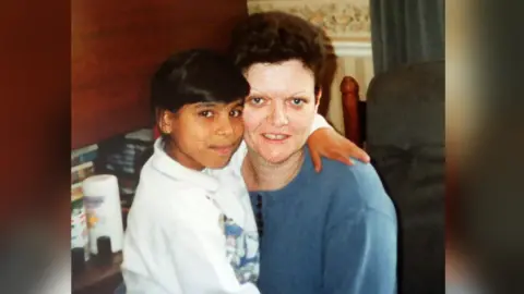 Skelton Family Philomena Skelton pictured hugging a child. Philomena has short dark brown hair, blue eyes and is wearing a blue top as she smiles at the camera.